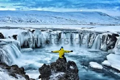Плакат Путешественник любуется величественным видом заснеженного водопада Годафосс (Исландия)